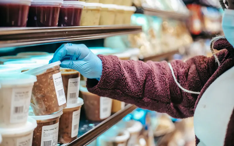 Gloved hand reaching for labeled deli containers stacked on a refrigerated store shelf